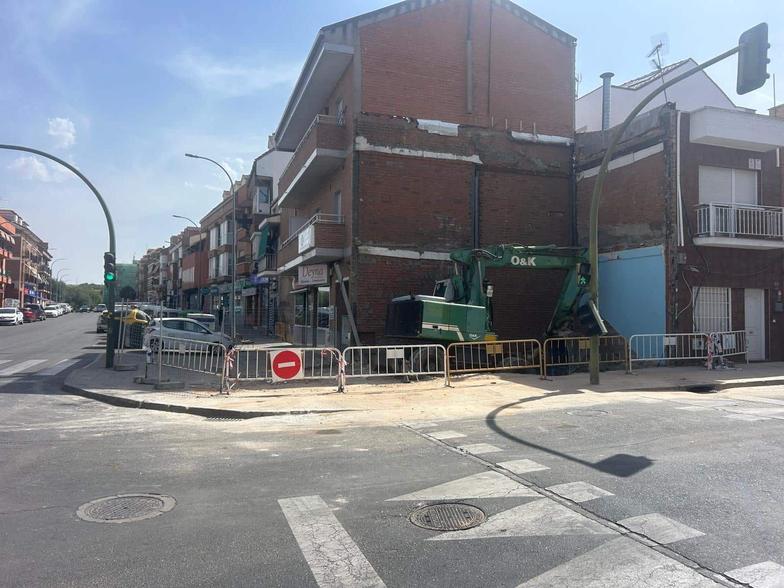 Sitio de construcción: edificio de ladrillo con excavadora en la esquina, rodeado de barreras y señal de prohibido el paso en un día soleado.