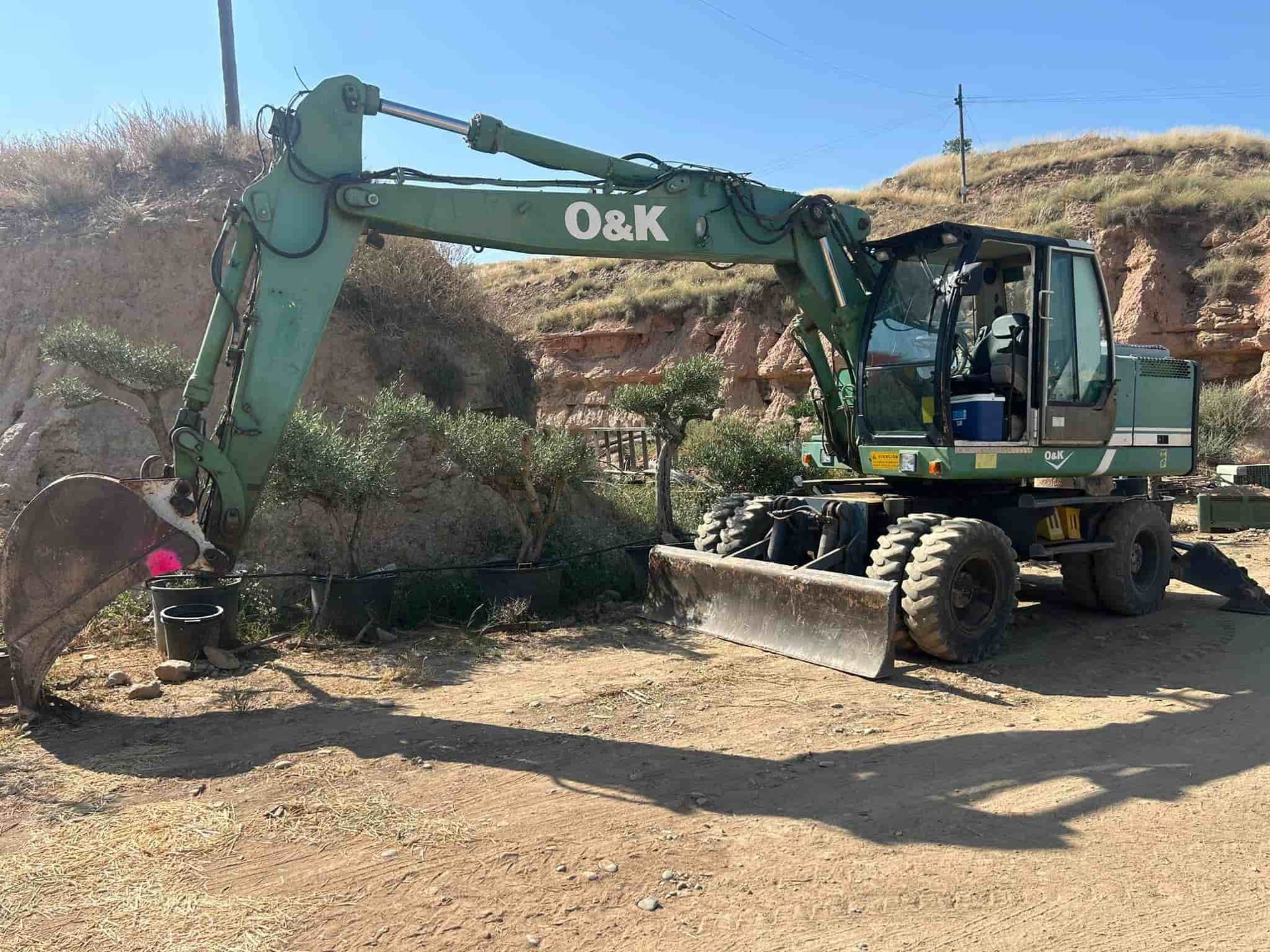 Excavadora O&K verde en un camino de tierra, con el cucharón hacia abajo y el brazo extendido cerca de una pequeña colina y plantas.