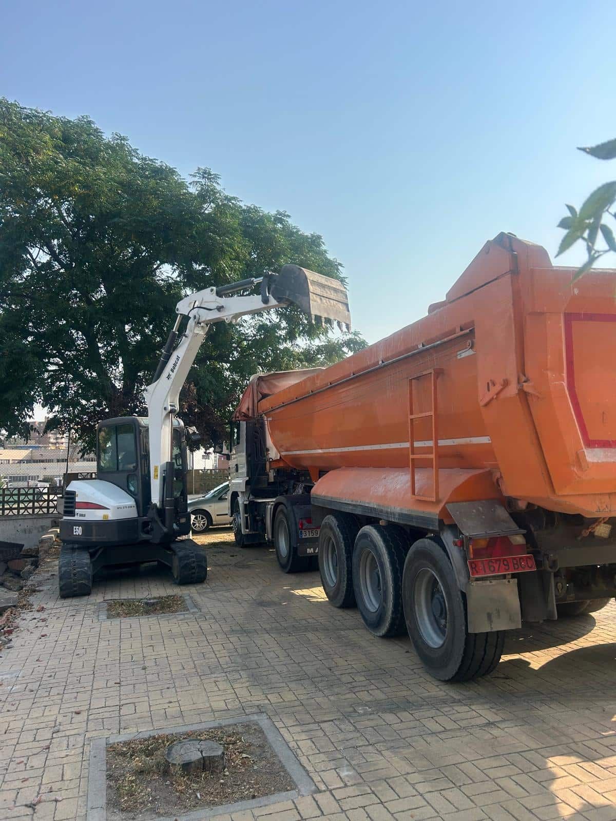 Excavadora Bobcat cargando un camión volquete naranja en una calle pavimentada con ladrillos; árbol en el fondo.