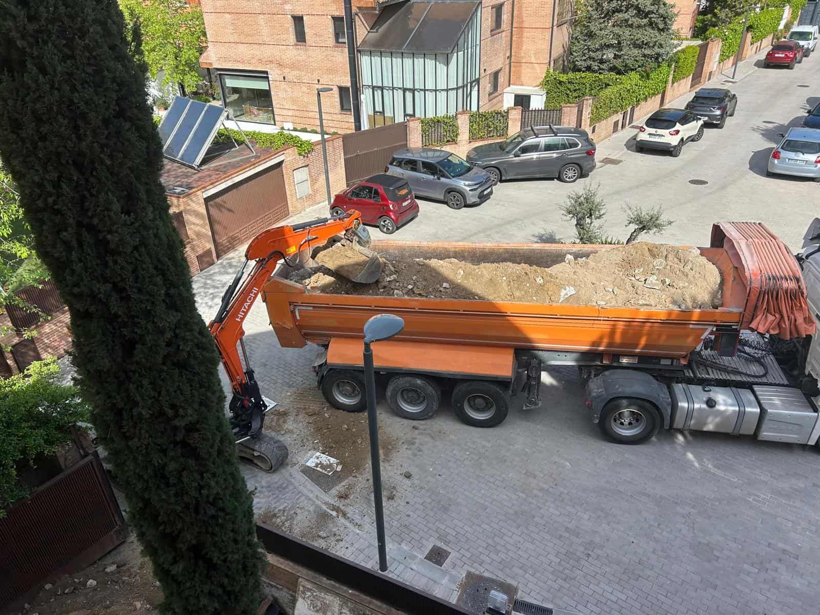 Un camión volquete naranja siendo llenado con tierra y rocas por una excavadora en una calle de la ciudad.