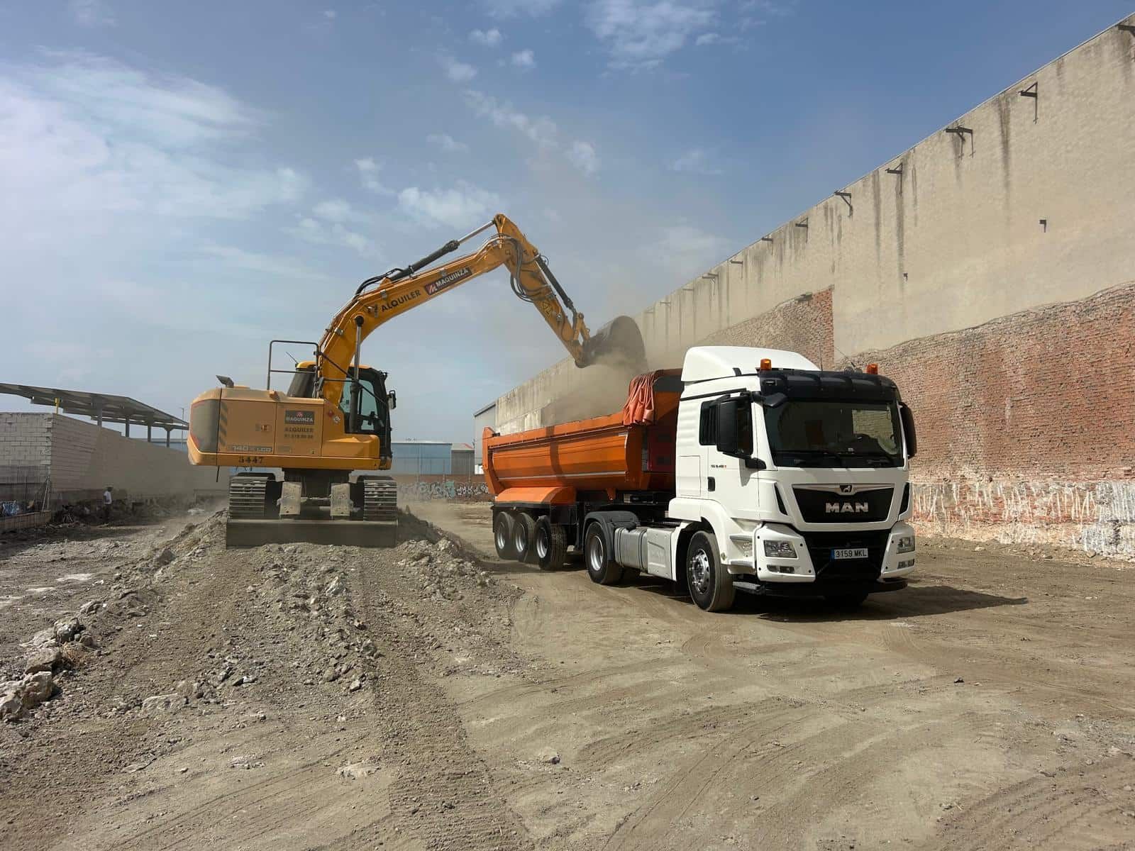 Excavadora amarilla cargando un camión volquete naranja con grava en un sitio de construcción.