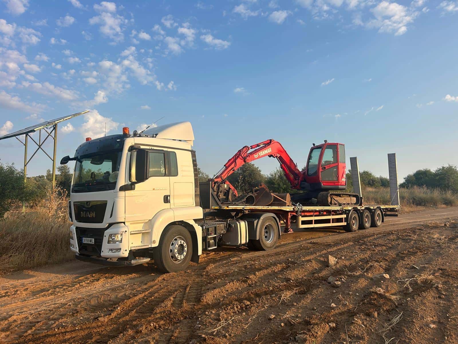 Camión MAN blanco que transporta una excavadora roja en un remolque de plataforma por un camino de tierra.