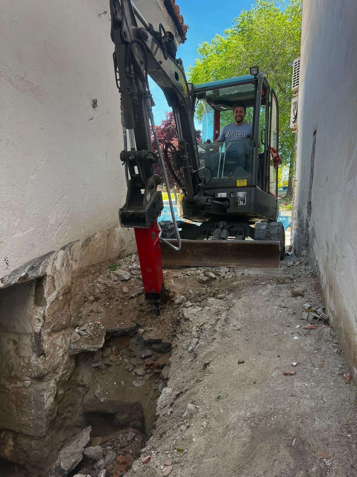 Hombre operando una miniexcavadora en un espacio estrecho, cavando cerca de un edificio.