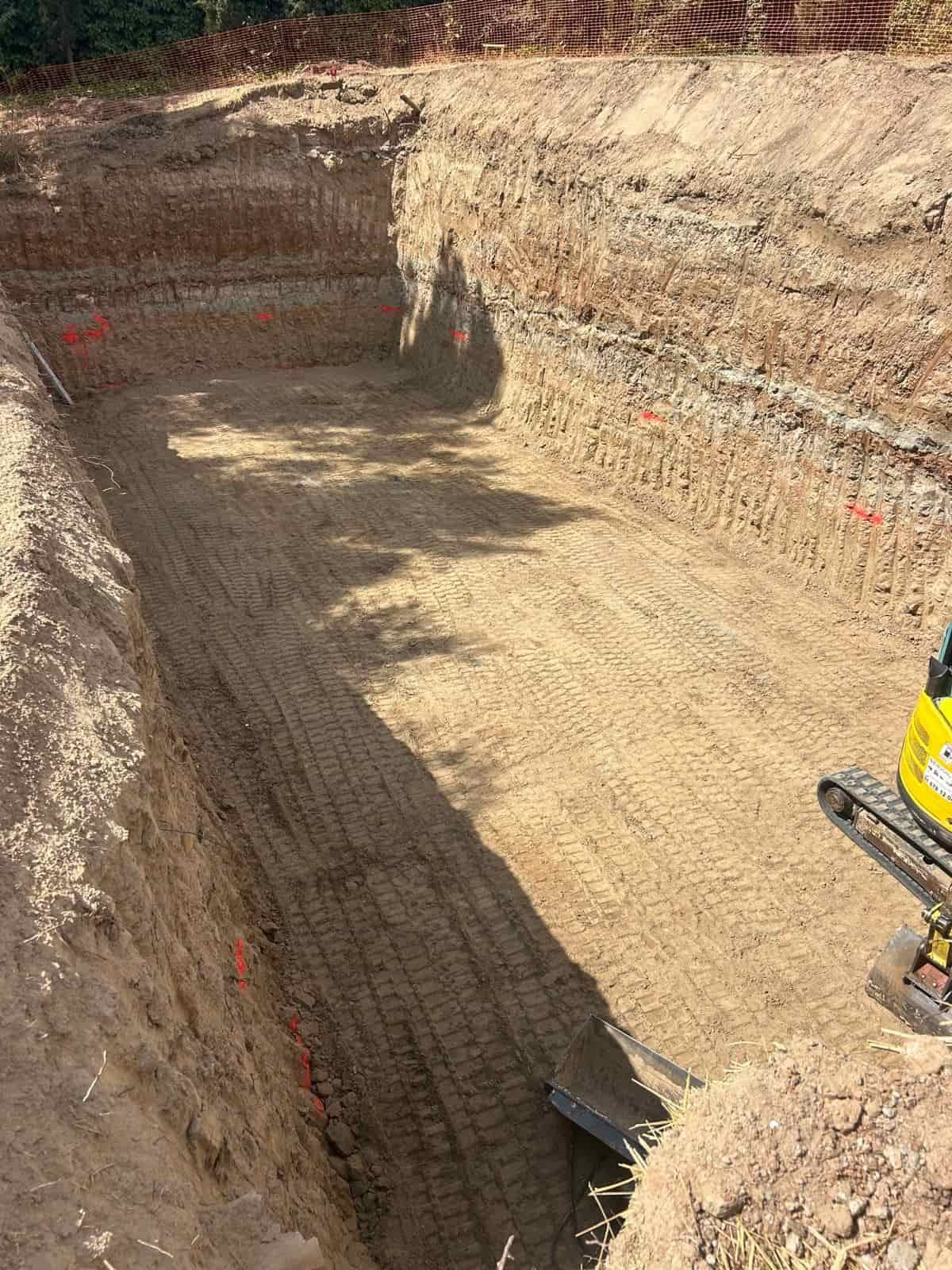 Solar excavado de forma rectangular, con muros escalonados de tierra arenosa y pequeña excavadora.