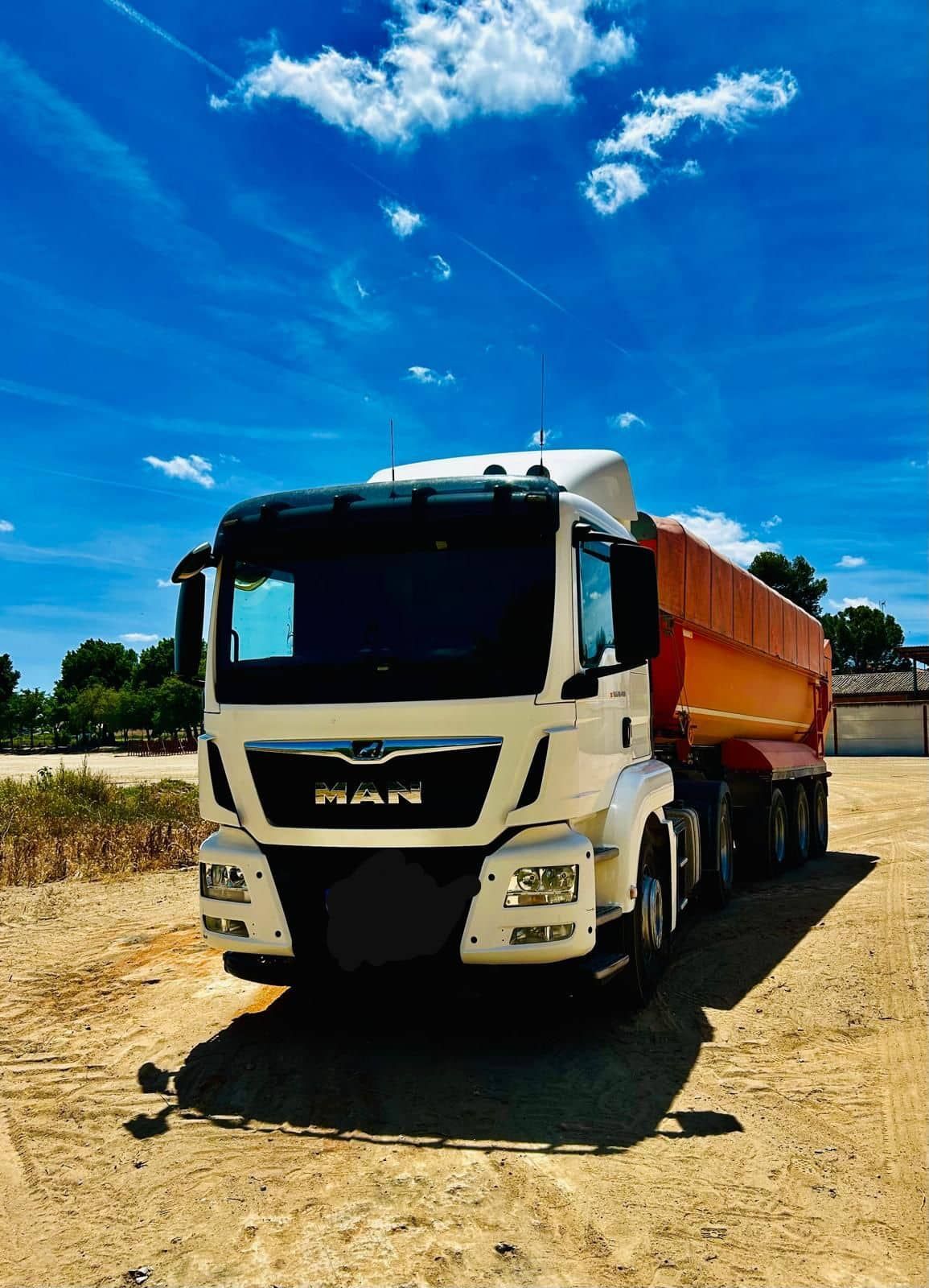 Camión MAN blanco con remolque naranja, estacionado sobre tierra bajo un cielo azul.