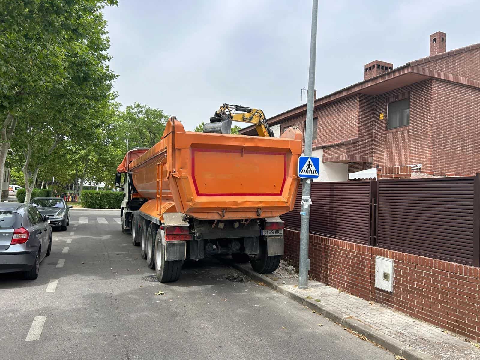 Camión volquete naranja estacionado en la calle con excavadora; cerca de una casa con cerca marrón.