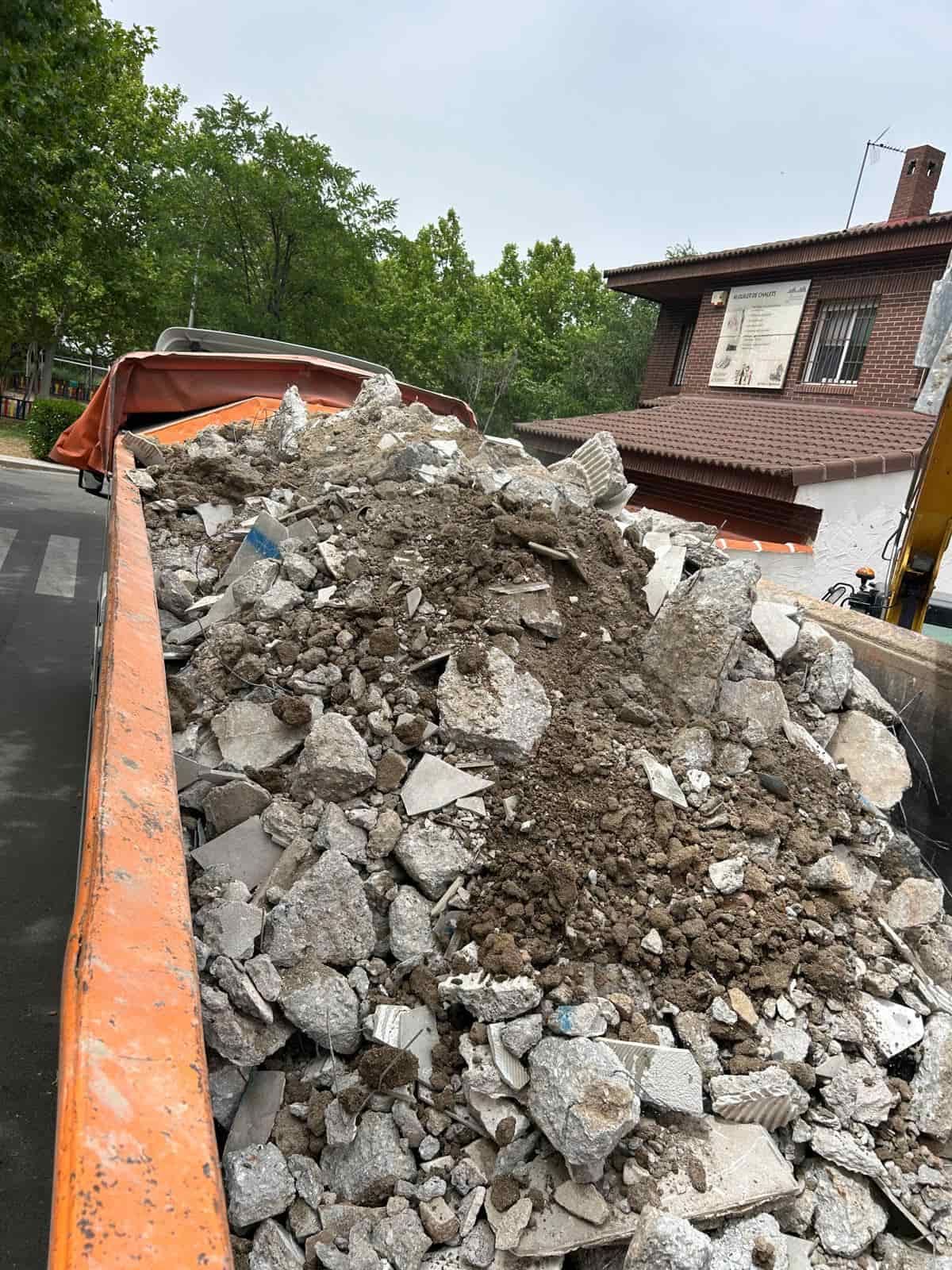 Una caja de camión rebosante de escombros, probablemente restos de demolición, frente a un edificio de ladrillos.