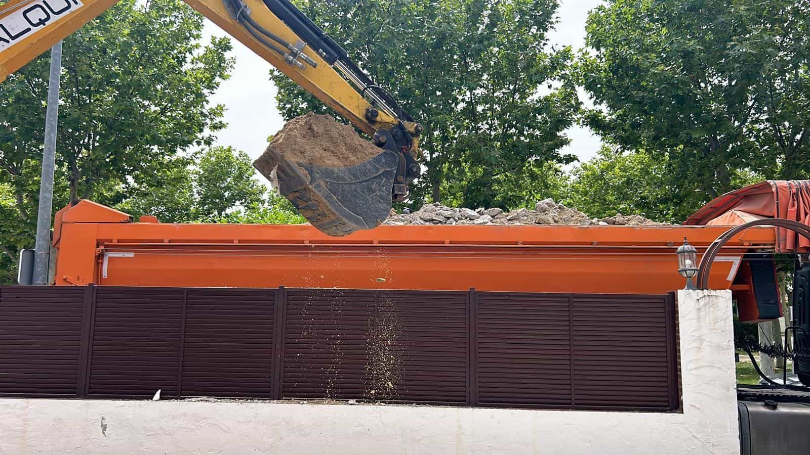Una excavadora arroja escombros en un camión volquete naranja, cerca de una valla marrón.