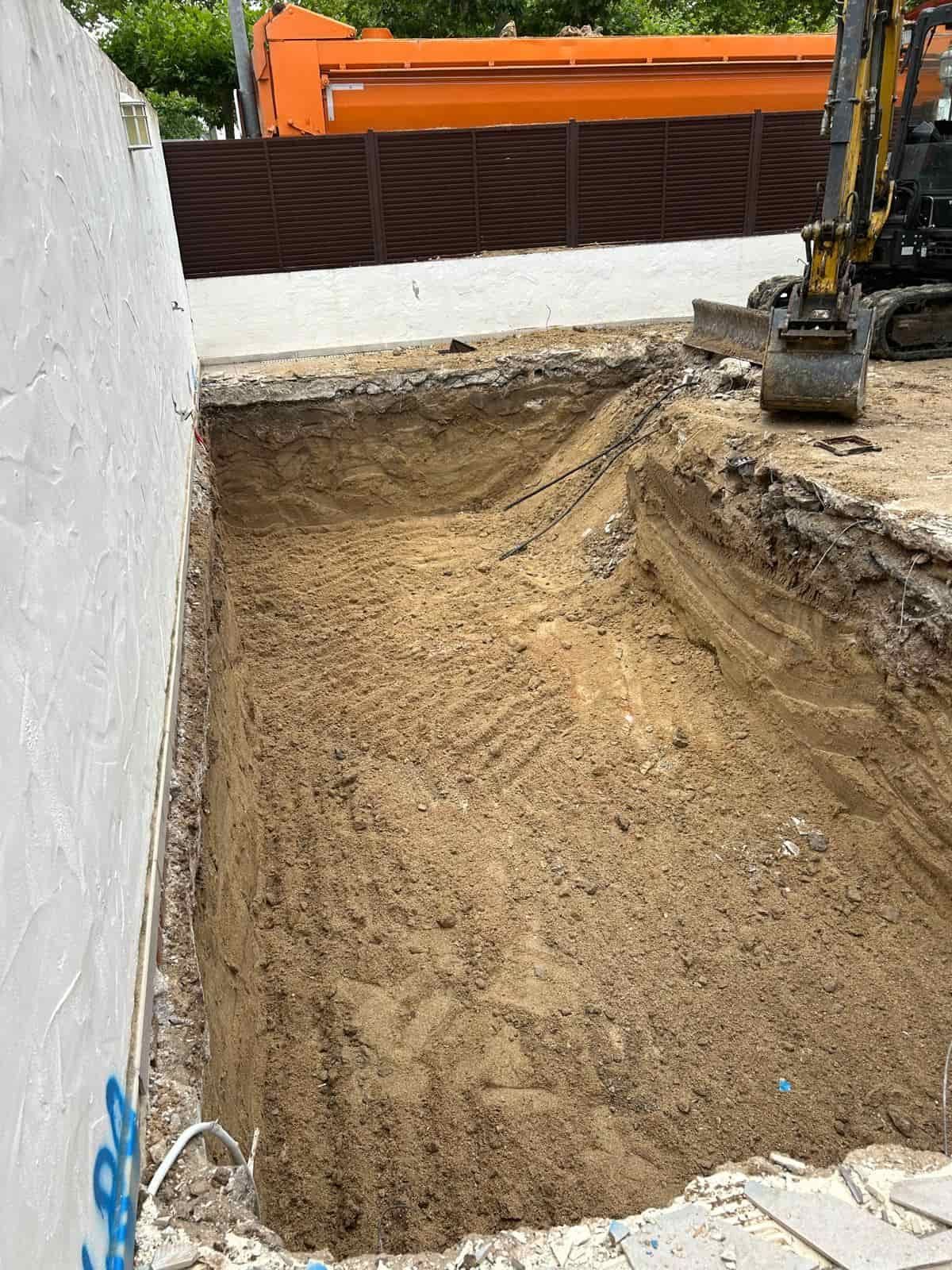 Una zanja rectangular cavada al lado de una pared blanca, equipo de construcción y un camión naranja.