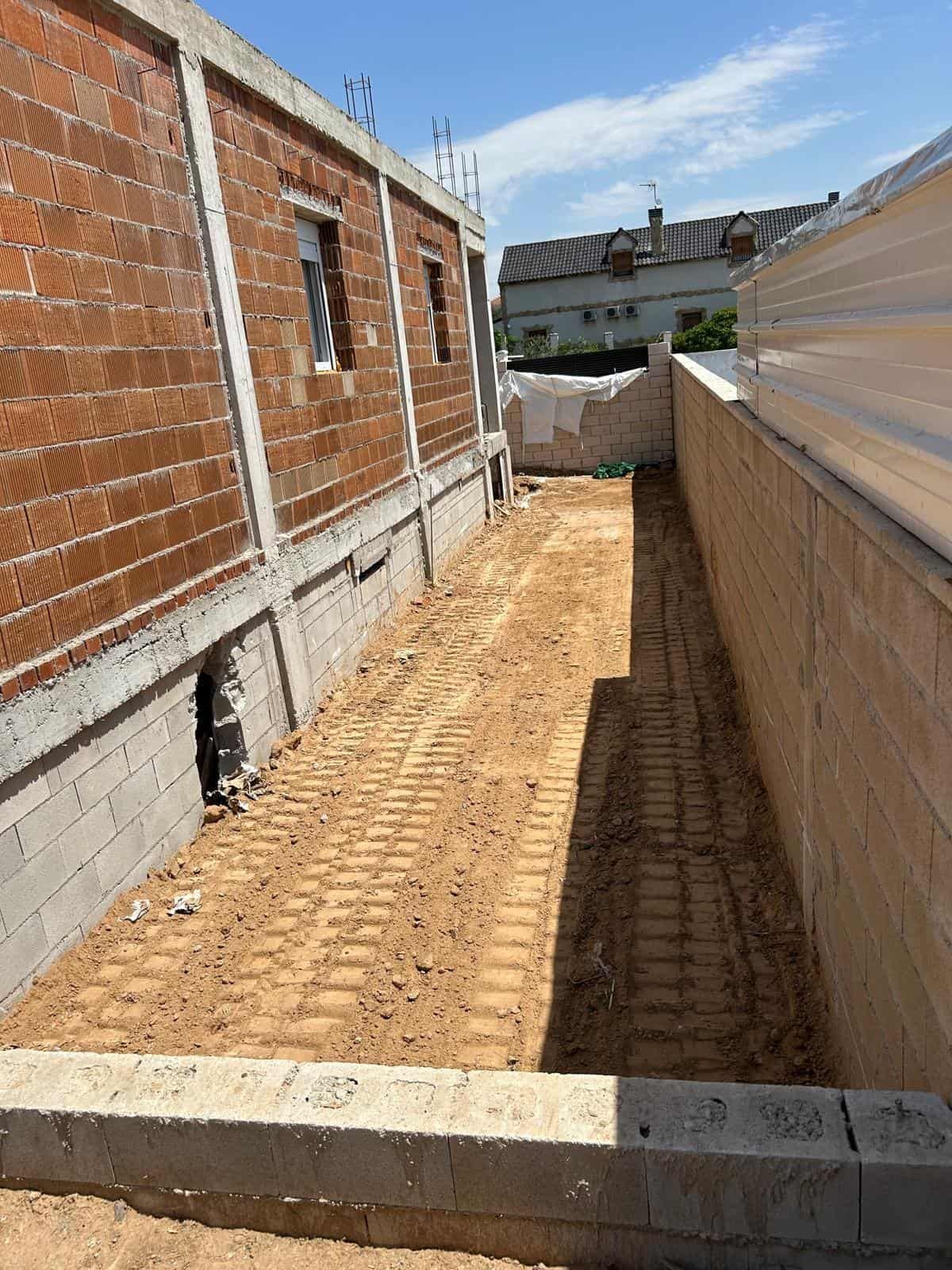 Obra de ladrillo y hormigón; estrecho sendero arenoso entre edificios. Cielo azul.