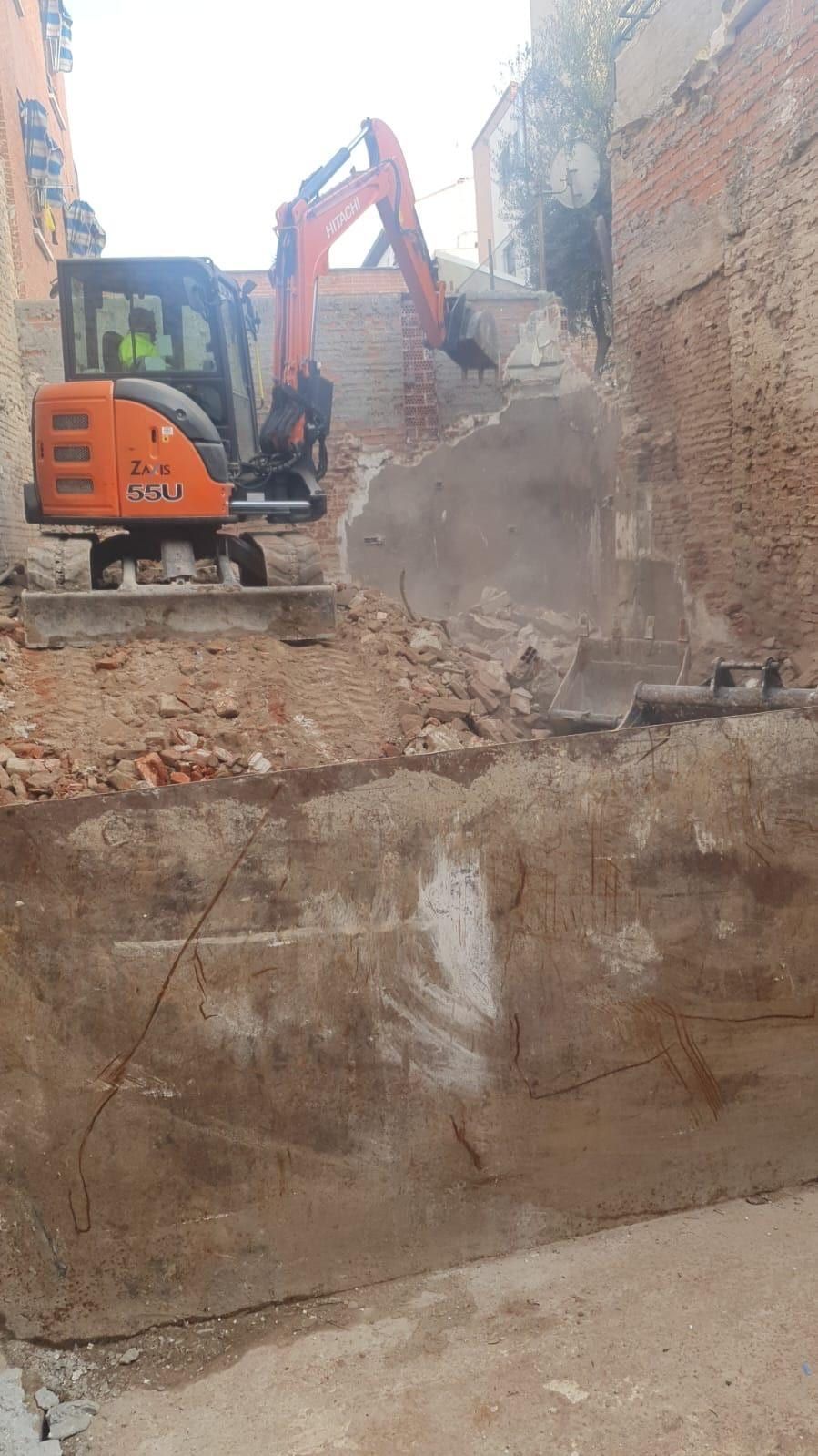 Excavadora demoliendo un muro de ladrillos en una obra. Máquina naranja, muro gris, luz natural.