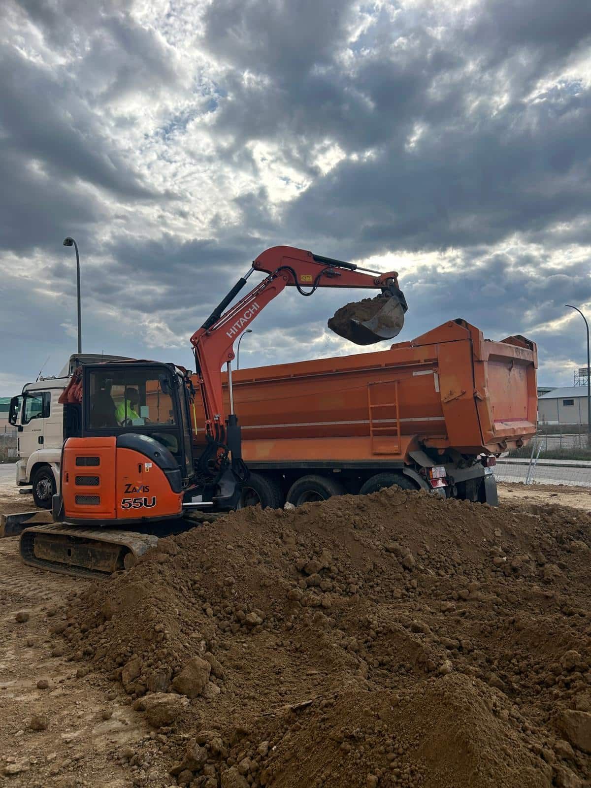 Excavadora naranja cargando tierra en un camión volquete naranja bajo un cielo nublado.