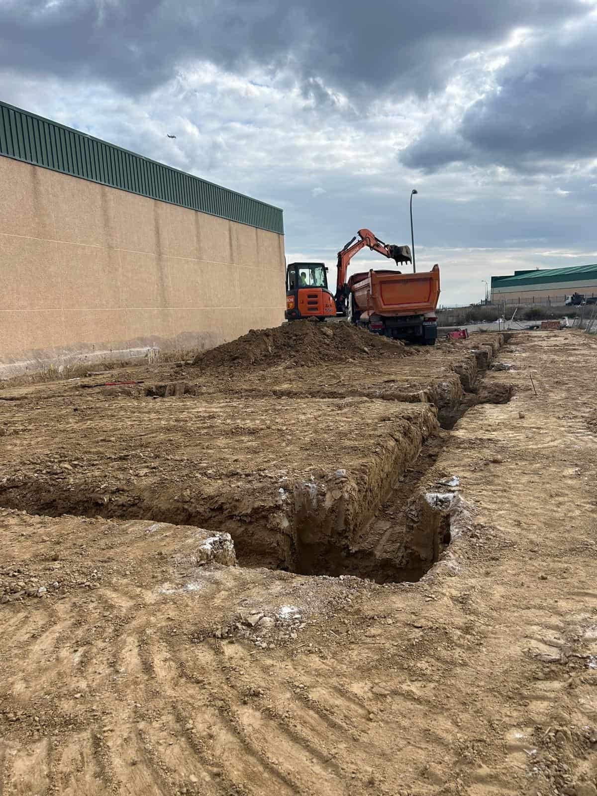 Obra con excavadora y camión volquete. Tierra excavada, edificio al fondo y cielo nublado.