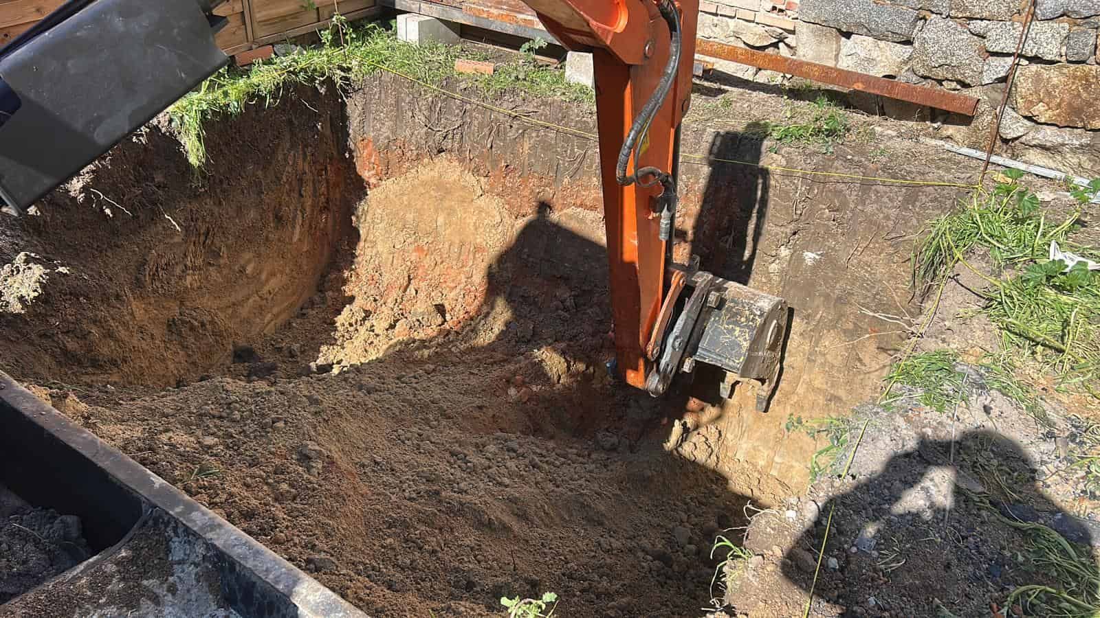Una excavadora naranja cavando un hoyo en el suelo, junto a un muro de piedra, en un entorno al aire libre.