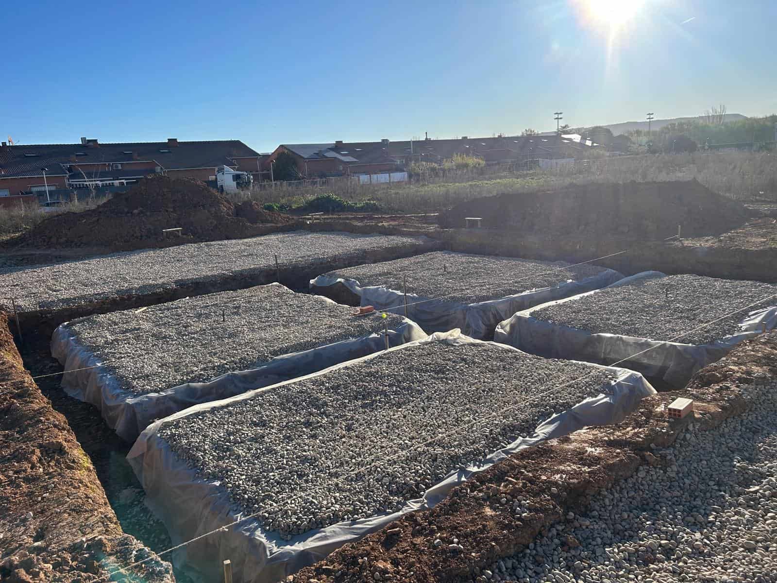 Terreno excavado con zonas rellenas de grava, cubierto con plástico, para un proyecto de construcción al aire libre bajo un cielo soleado.