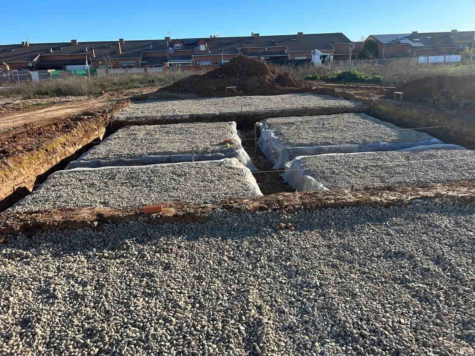 Zanjas rellenas de grava en un sitio de construcción, preparadas para una base, con un montículo de tierra y casas al fondo.