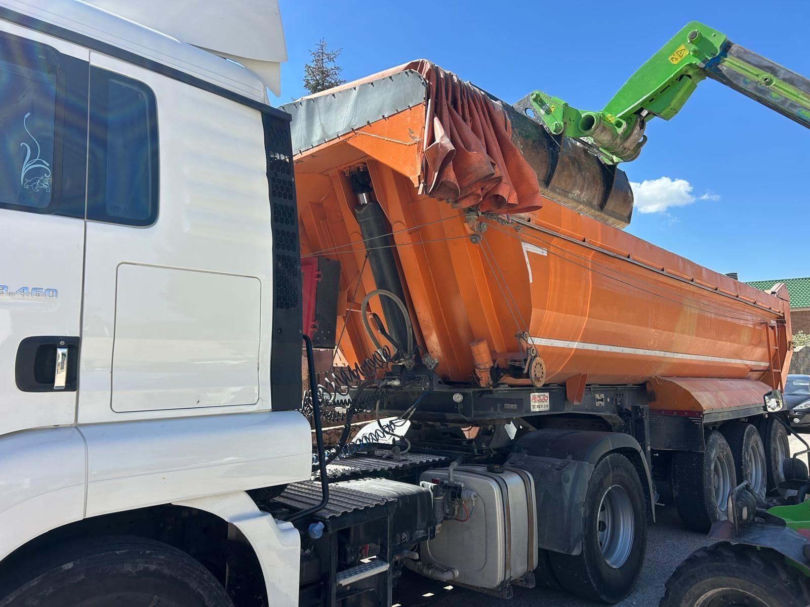 Un camión volquete naranja siendo cargado por una máquina verde bajo un cielo soleado.
