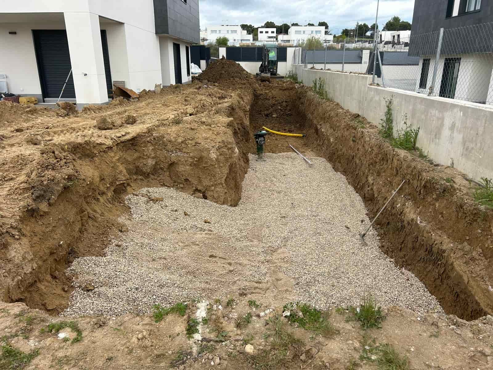 Zanja excavada rellena de grava, sitio de construcción cerca de casas modernas.