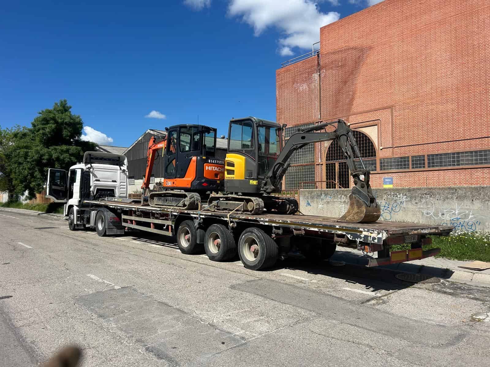 Un camión de plataforma transporta dos excavadoras en una calle de la ciudad frente a un edificio de ladrillos.
