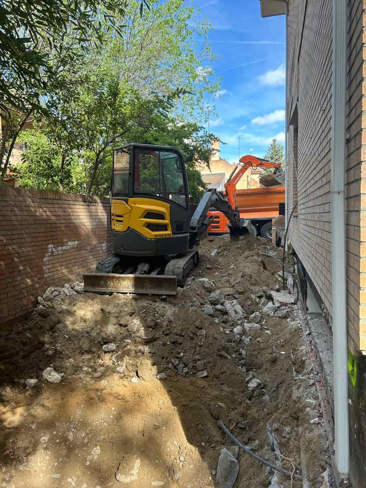 Excavadora amarilla cavando junto a un edificio de ladrillos, con un camión naranja al fondo.