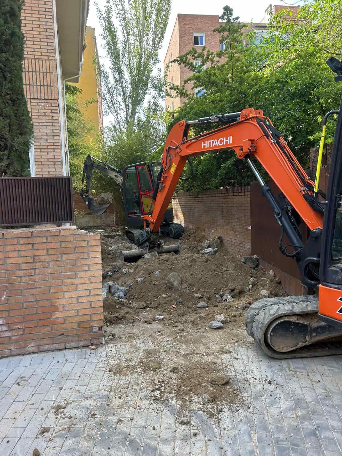 Dos excavadoras Hitachi de color naranja excavando en un sitio de construcción al aire libre con una pared de ladrillos y árboles.