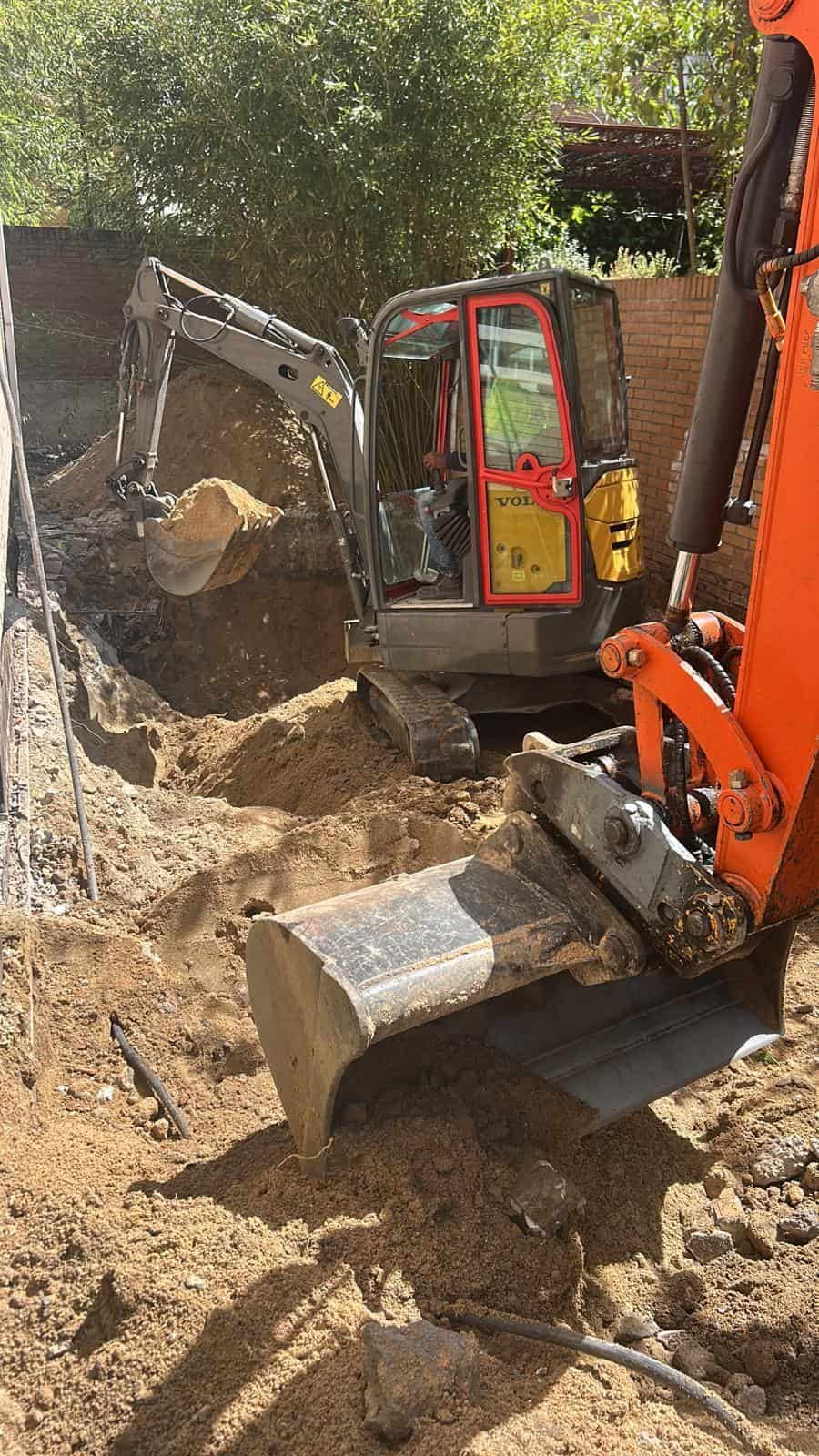 Miniexcavadora cavando en una zanja junto a una excavadora naranja, día soleado.