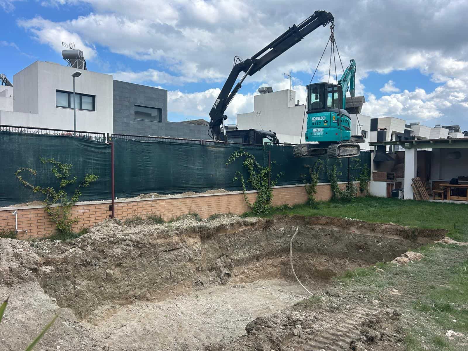 Excavadora cavando un hoyo rectangular en un patio detrás de una valla y edificios blancos bajo un cielo azul.