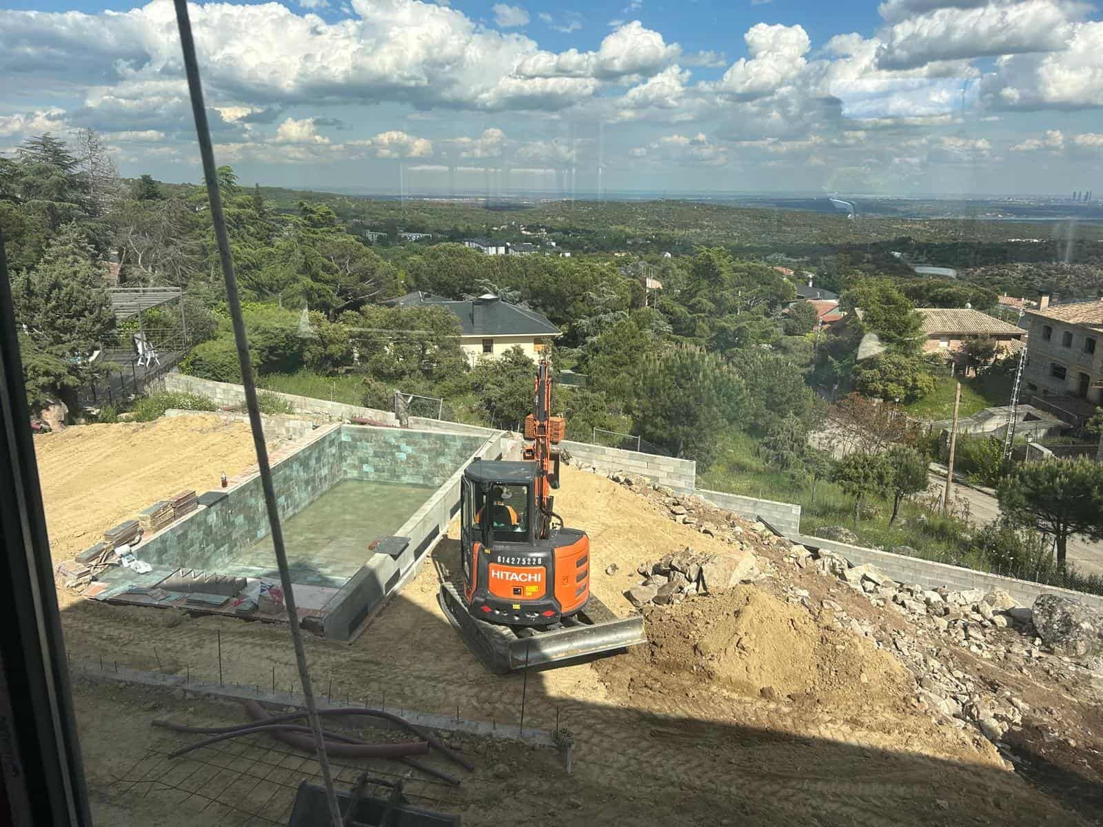 Sitio de construcción con una excavadora naranja excavando cerca del marco de una piscina de hormigón, con vista a un valle.