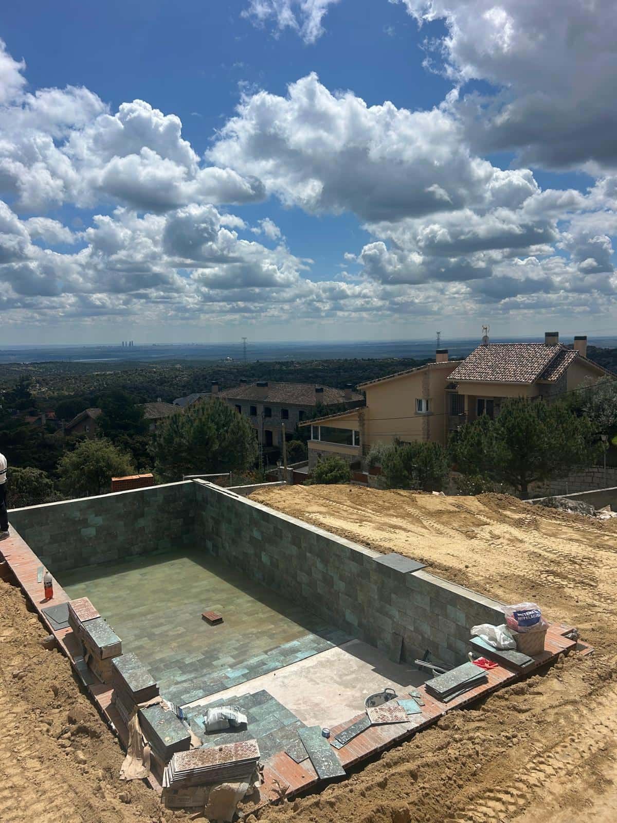 Piscina en construcción con vista a las casas y al paisaje lejano bajo un cielo nublado.