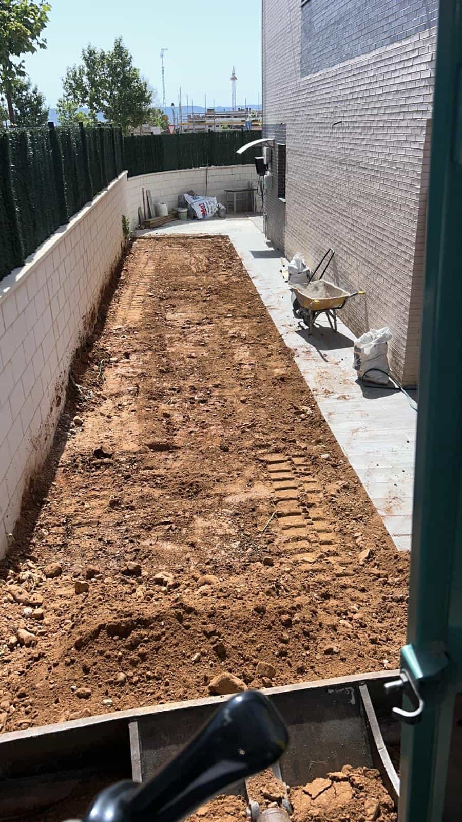 Patio trasero largo y estrecho en preparación para jardinería. Tierra marrón, paredes blancas, un lateral de casa y una carretilla.
