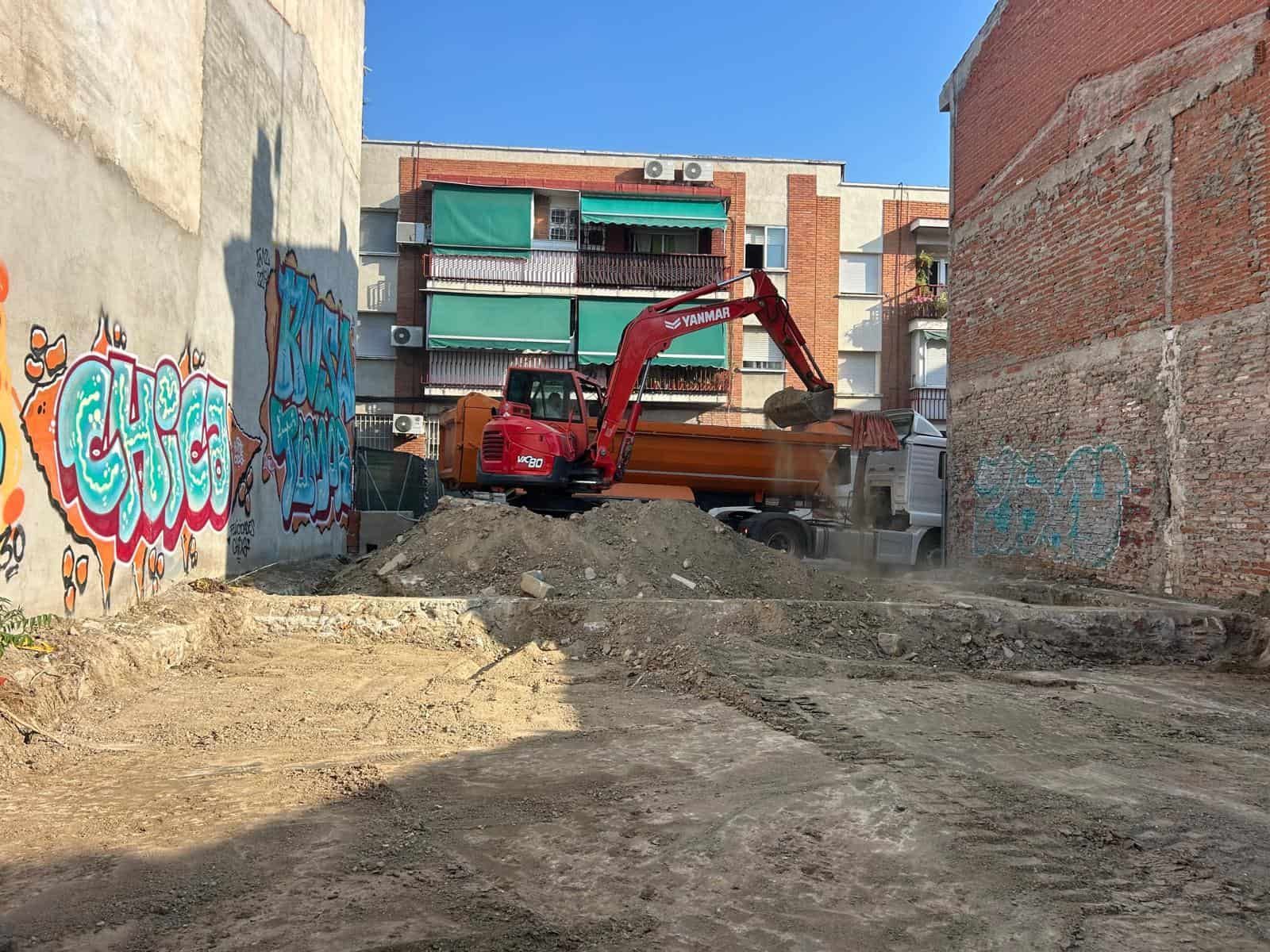 Una excavadora roja cargando tierra en un camión volquete en un sitio de construcción entre edificios; grafitis en las paredes.