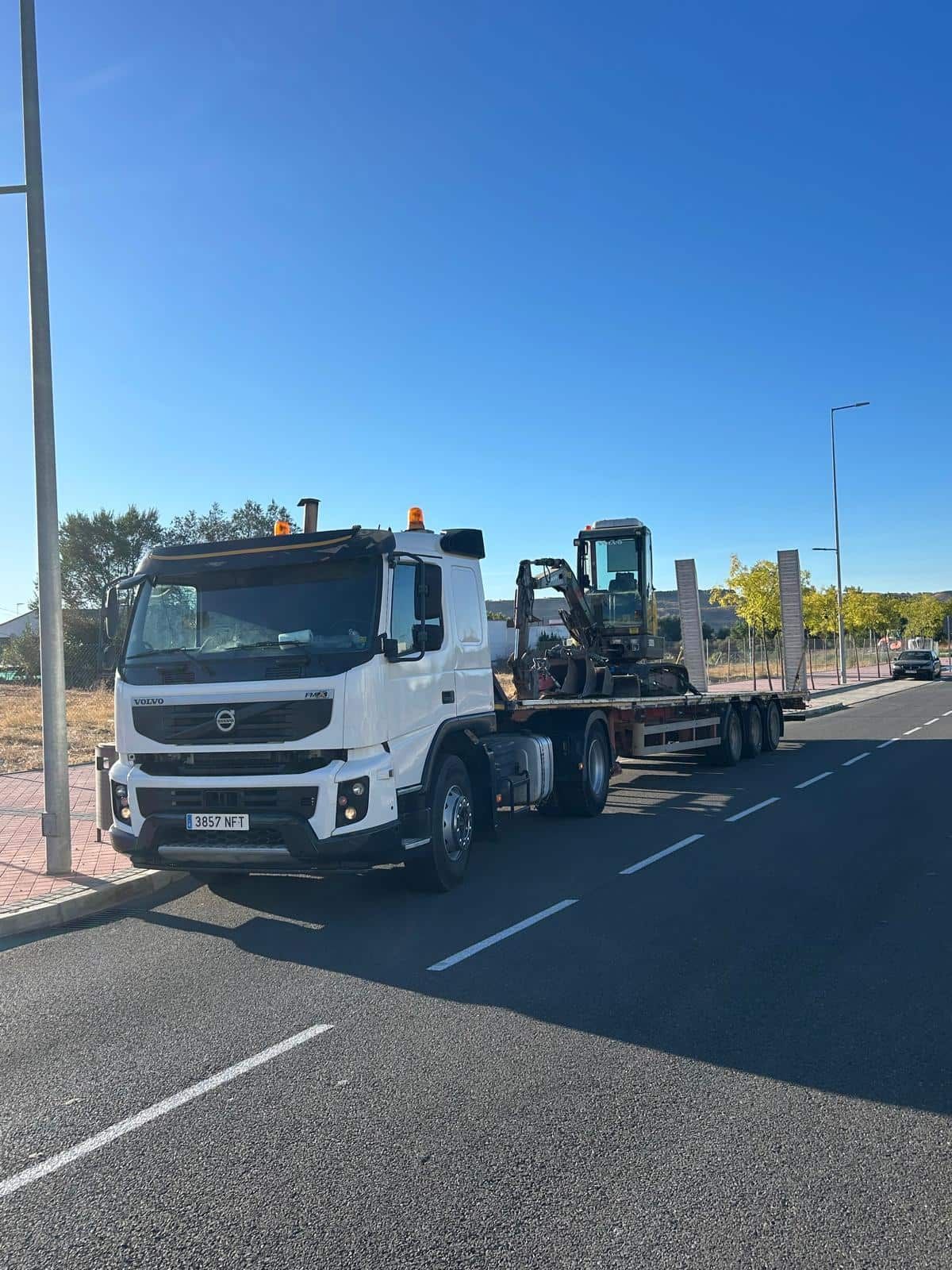Camión blanco con equipo de construcción sobre un remolque de plataforma estacionado en una carretera en un día soleado.