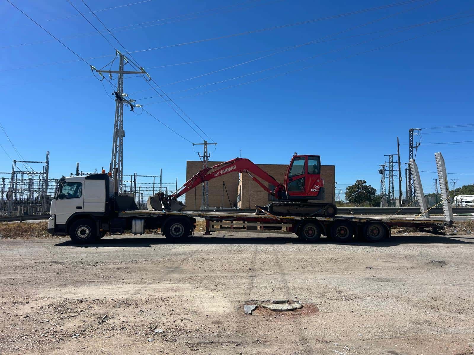 Camión que transporta una excavadora roja en un remolque de plataforma cerca de líneas eléctricas y una subestación bajo un cielo azul.