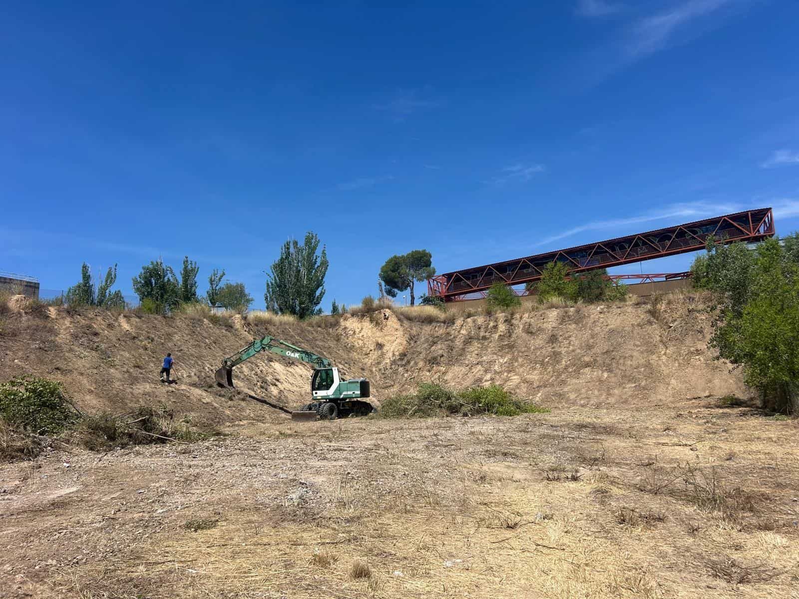Excavadora limpiando terreno seco cerca de un puente oxidado bajo un cielo azul claro.