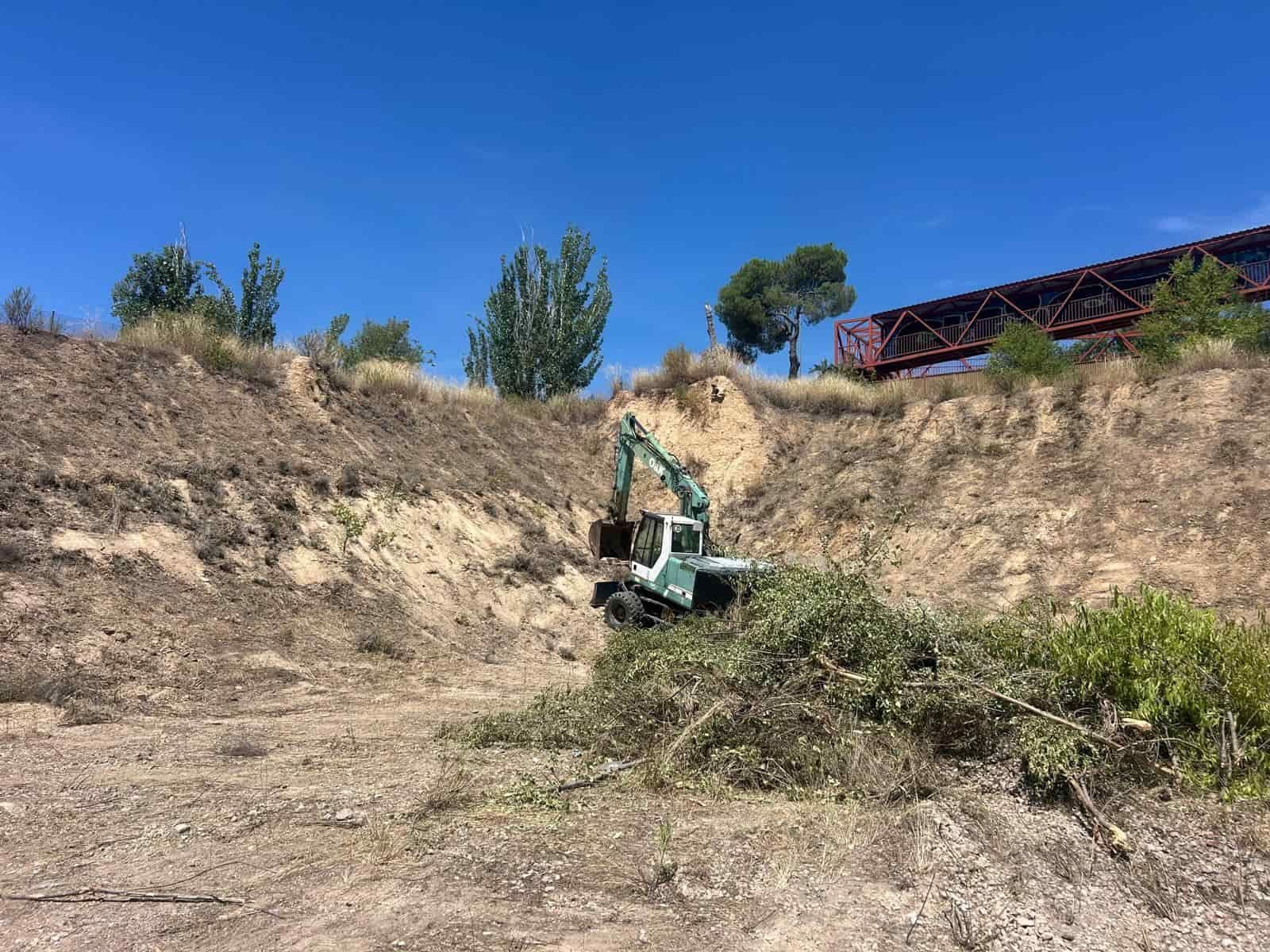 Una excavadora quitando maleza de una ladera bajo un cielo azul, cerca de un puente.