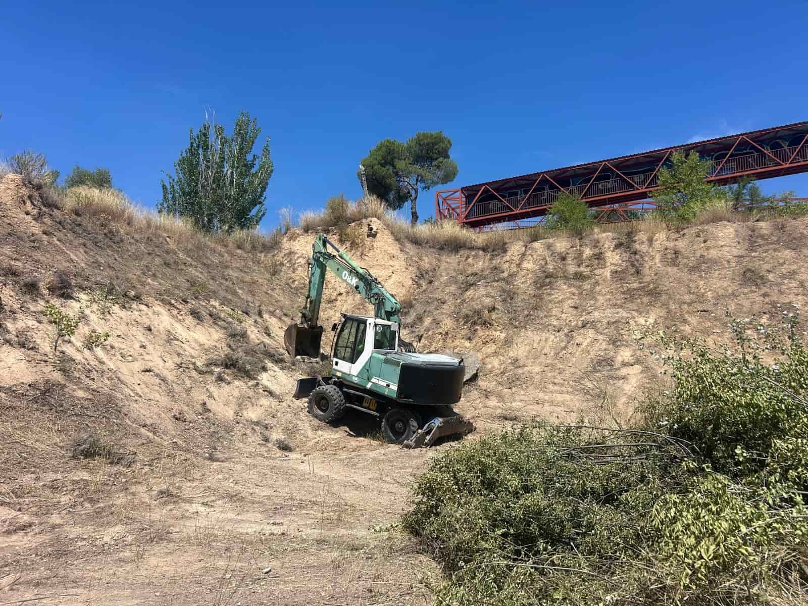 Una excavadora en una zanja seca, debajo de un puente de metal rojo, en un día soleado.