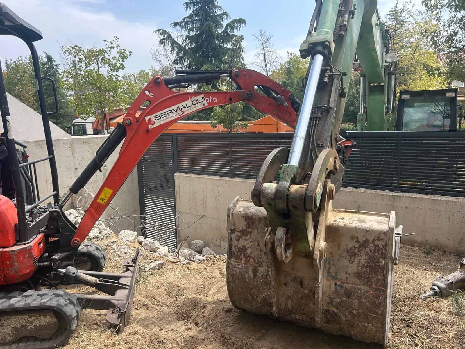 Una pequeña excavadora roja con un cucharón cerca de una pared, utilizada para construcción o excavación al aire libre.