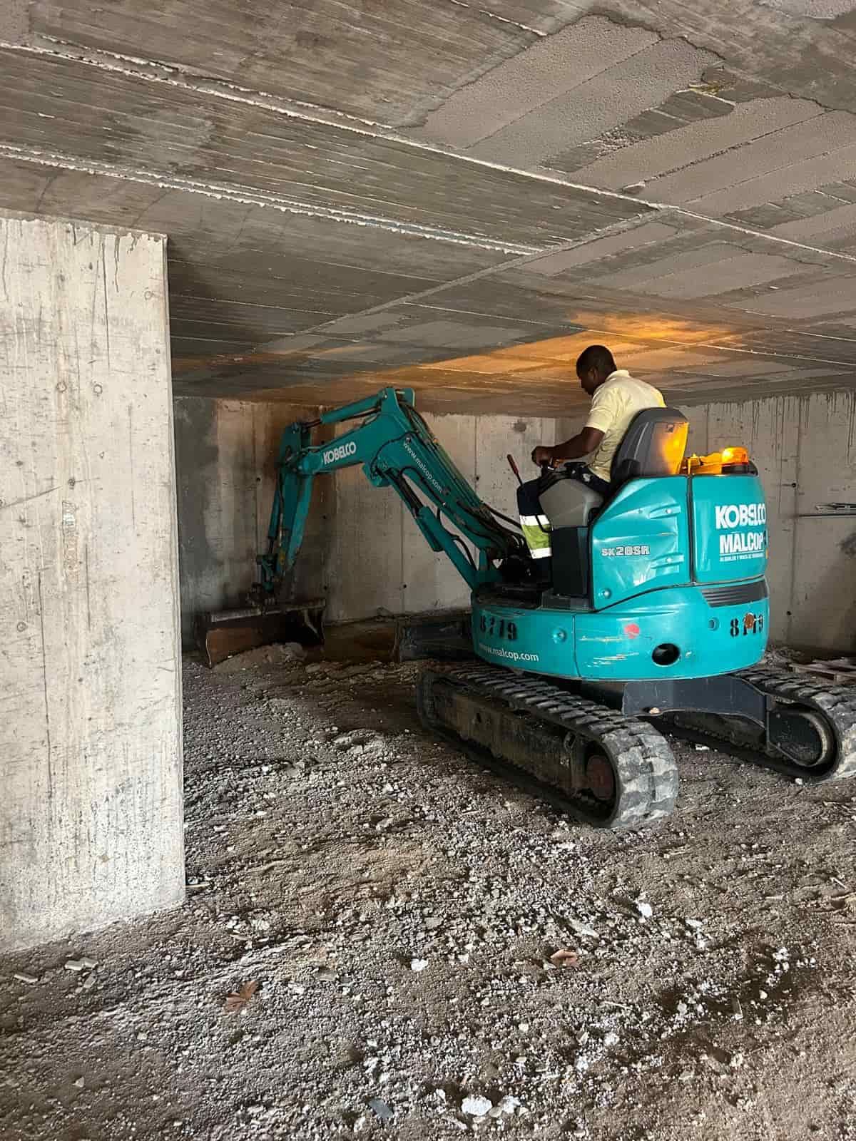 Un hombre opera una excavadora debajo de una estructura de concreto, limpiando escombros.