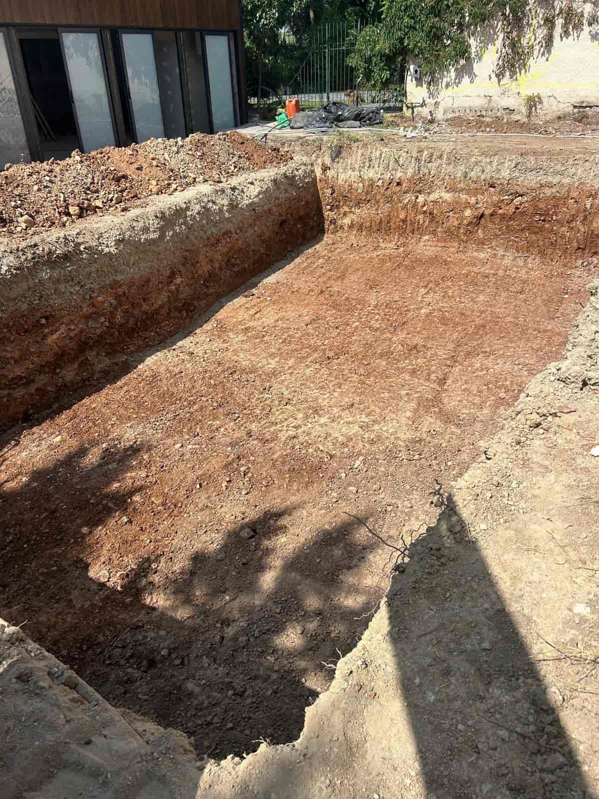 Un hoyo de excavación rectangular en suelo de color marrón rojizo, presumiblemente para construcción, al aire libre y bajo la luz del sol.