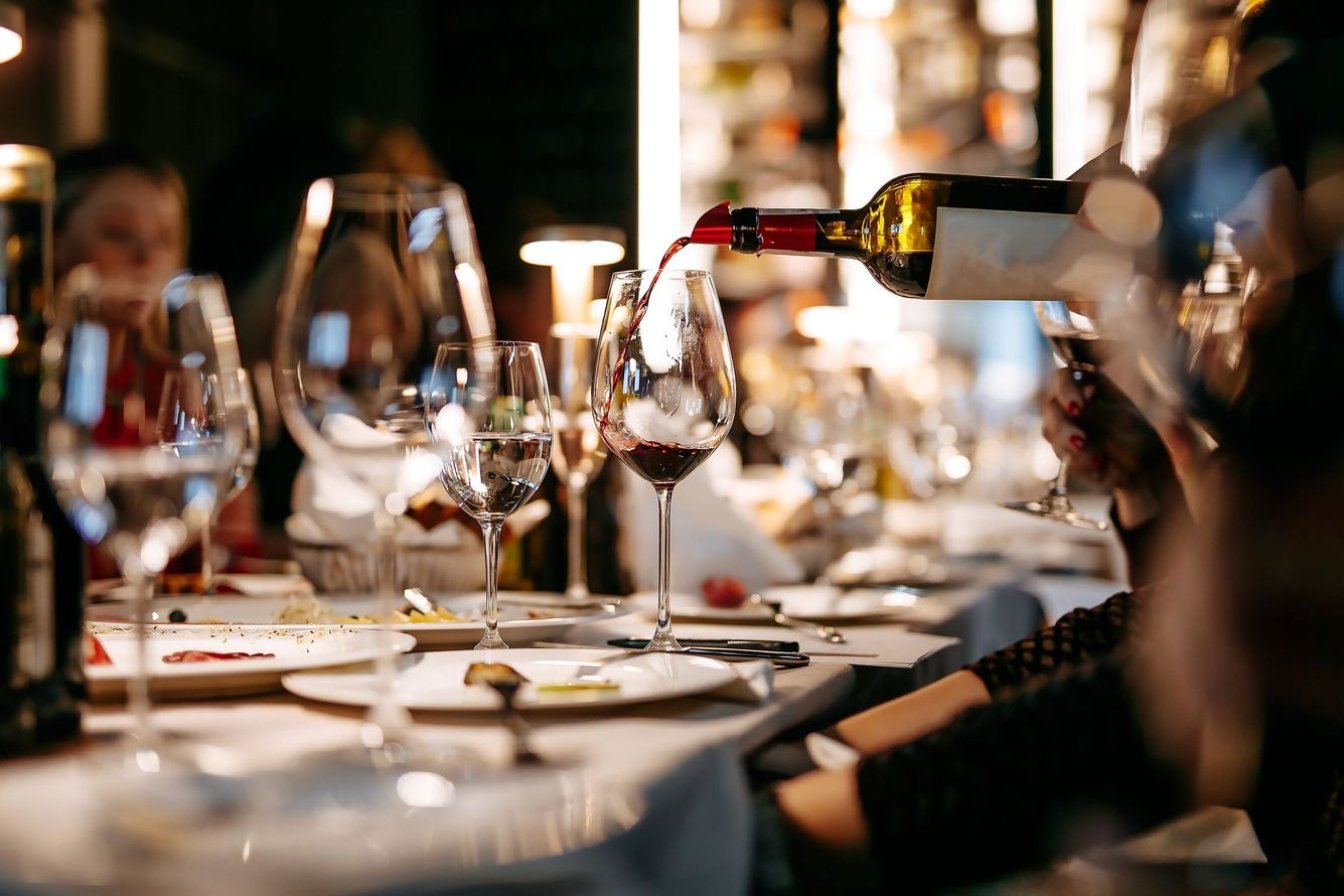 Du vin est versé dans un verre sur une table à dîner faiblement éclairée, avec plusieurs verres à vin et couverts disposés.