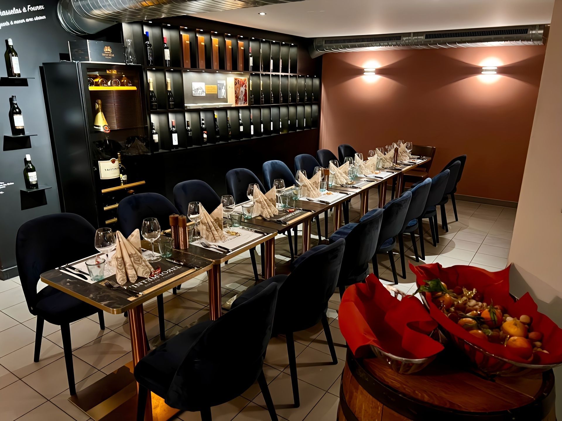 Salle à manger élégante avec une longue table éclairée aux chandelles, des chaises noires et des verres à vin devant un mur sombre.