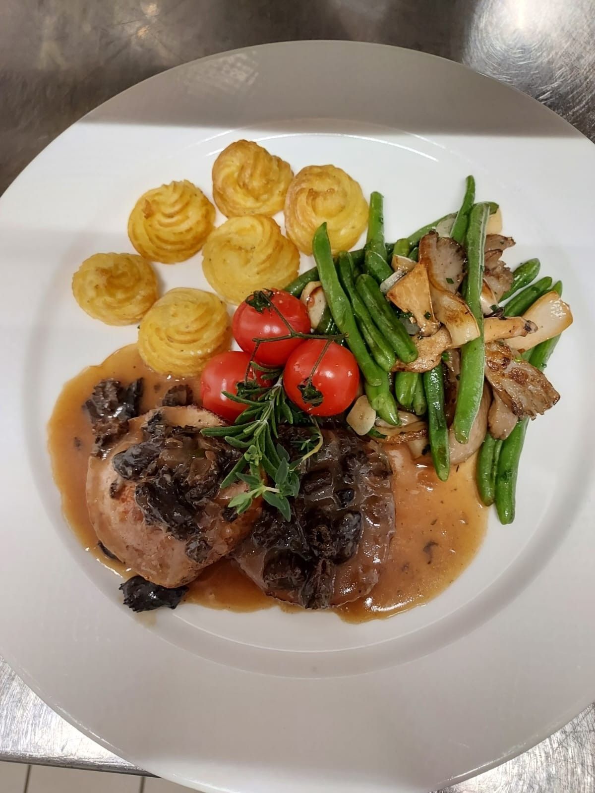 Plat gastronomique servi à l'assiette, composé de viande rôtie, de purée de pommes de terre, de haricots verts, de tomates cerises et de champignons.