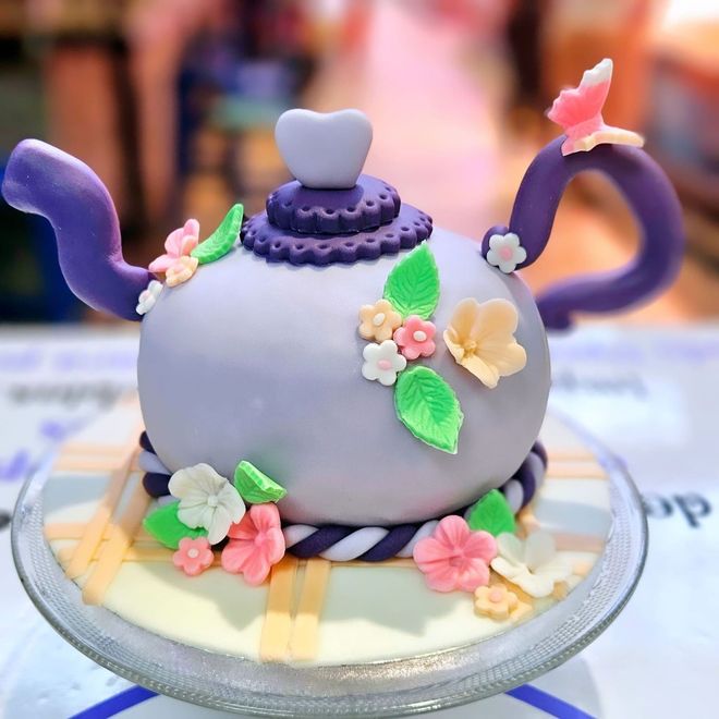 Un pastel morado en forma de tetera decorado con flores y mariposas.