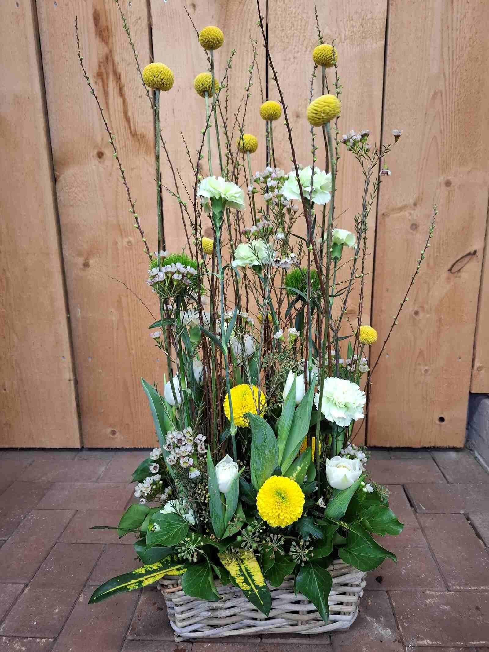 Ein Korb voller gelber und weißer Blumen steht auf einem Ziegelboden vor einer Holzwand.