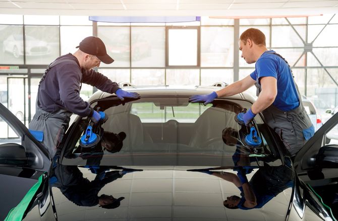 Zwei Automechaniker ersetzen in einer Werkstatt die Windschutzscheibe eines schwarzen Autos. Sie tragen Handschuhe und benutzen Werkzeuge.