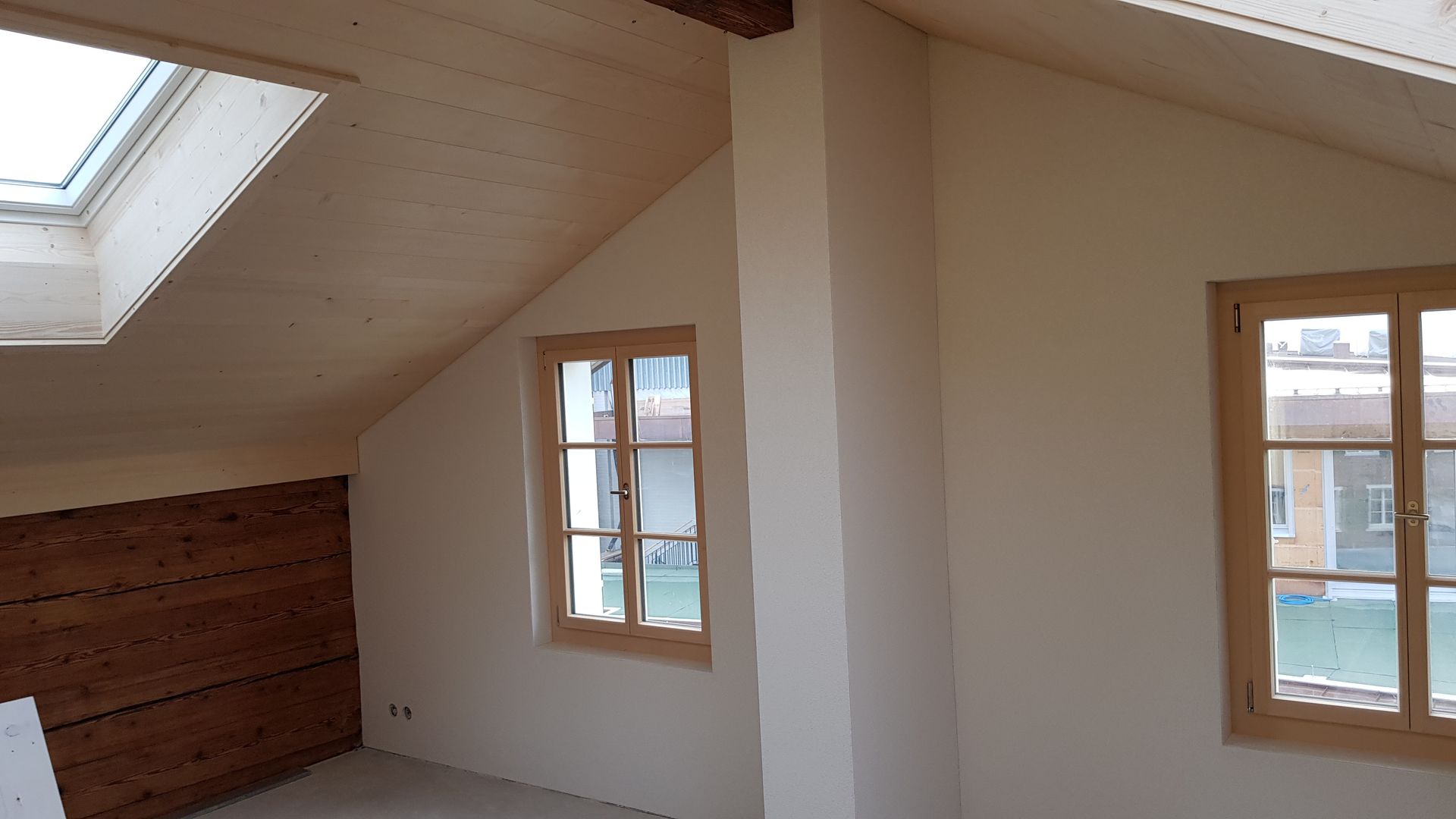 Ein Kellerraum mit freiliegenden Holzbalken an der Decke, Dämmung, verputzten Wänden, zwei Fenstern und einem Heizkörper.