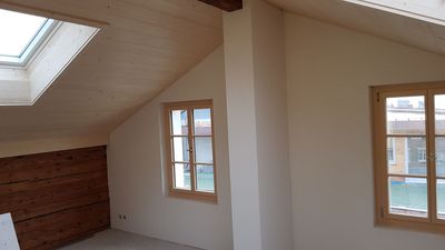 Ein Kellerraum mit freiliegenden Holzbalken an der Decke, Dämmung, verputzten Wänden, zwei Fenstern und einem Heizkörper.