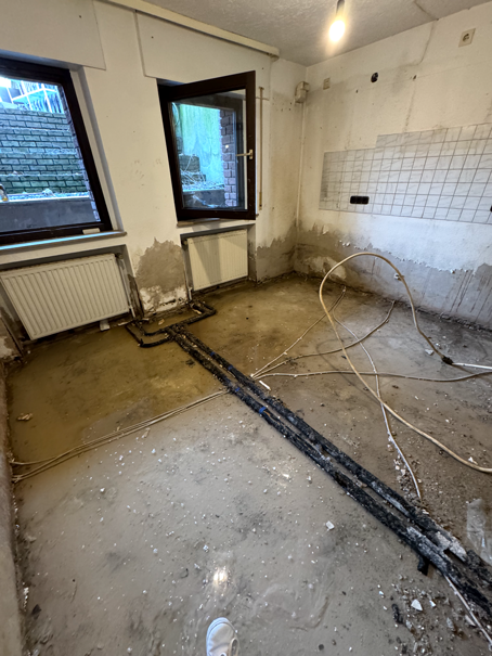 Teilweise renoviertes Zimmer mit blankem Betonboden, freiliegenden Leitungen und unfertigen Wänden, mit zwei Fenstern.