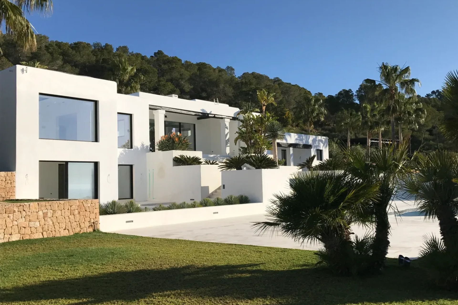 Villa moderna blanca con grandes ventanales y muro de piedra, en una ladera soleada.