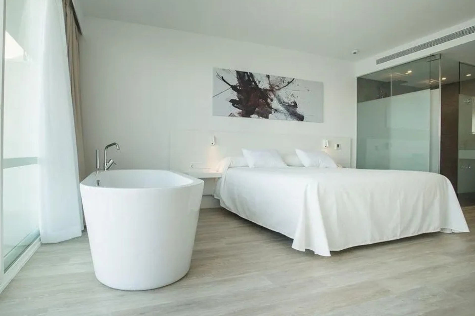 Dormitorio de hotel moderno con bañera blanca y cama tamaño king, con arte abstracto y baño con mampara de cristal.